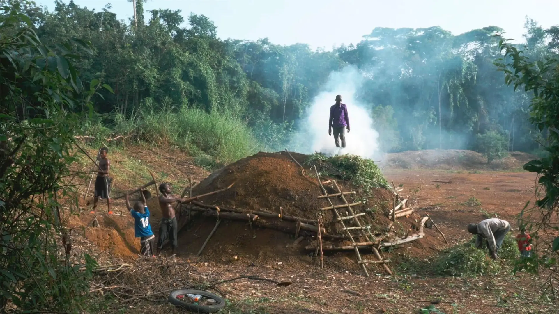 Charcoal burners working a smoking earth mound at the forest edge