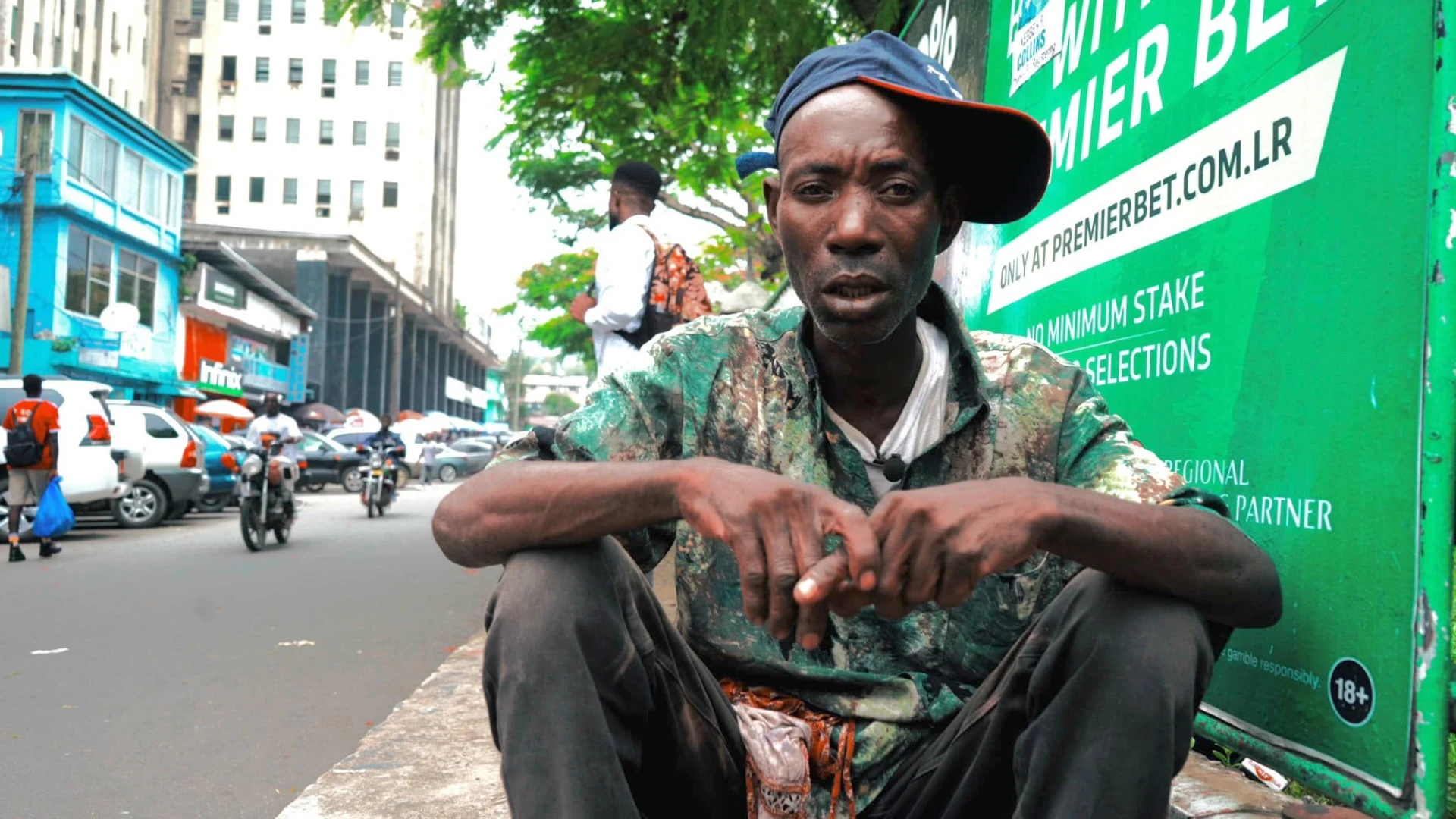 Man sitting on a curb speaking to camera in a busy city street